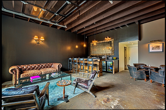 A dark lounge featuring leather sofas, armchairs, a small coffee table, and a bar in the background.