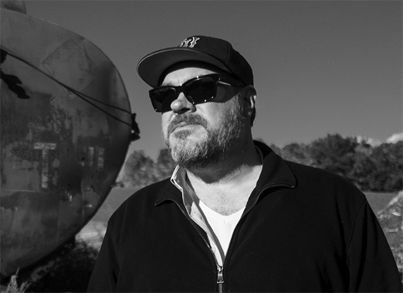 Black and white portrait of a bearded man wearing a baseball cap and sunglasses outdoors.