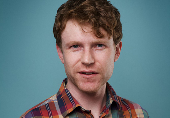 A man with wavy reddish-brown hair and a plaid shirt looks forward against a solid blue background.