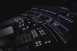 Close-up view of a dark professional audio mixing console with illuminated buttons, knobs, and faders.