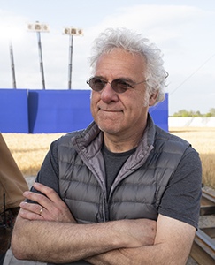 A man with curly grey hair and glasses wearing a grey puffer vest stands with arms crossed.