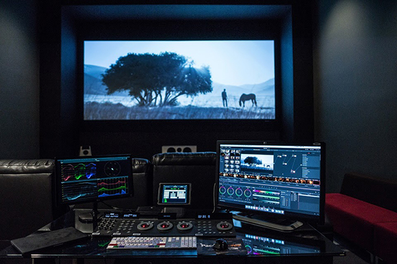A dark editing suite featuring a large projection screen showing a landscape and multiple computer monitors.