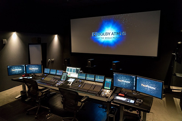A dark professional audio mixing suite with multiple monitors, large mixing consoles, and a projection screen.