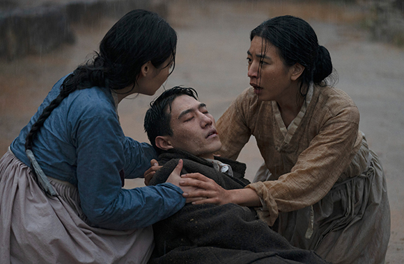 Two women in traditional Korean clothing support a man lying on the ground in the rain.