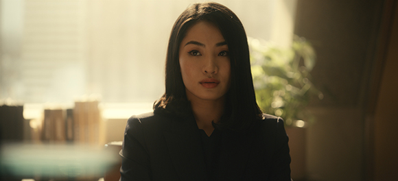 A woman with dark hair and a dark blazer looks forward in a softly lit room.