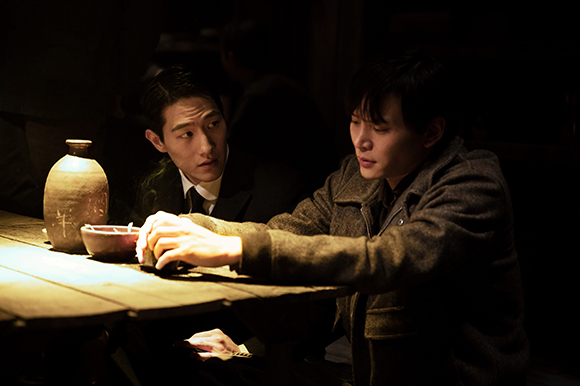 Two men sit at a wooden table in a dark room, one leaning forward while looking at the other.
