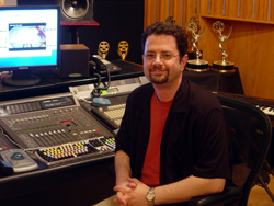 A man with glasses and a goatee sits behind a large audio mixing desk in a recording studio.