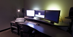 A dark workspace featuring three computer monitors, a keyboard, a mixing console, and studio speakers.