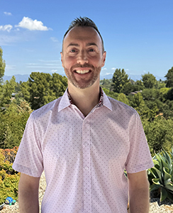 A smiling man wearing a light pink short-sleeved button-down shirt against a background of green trees.