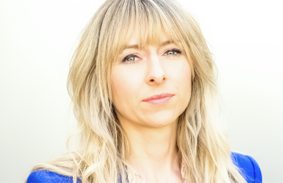 Close-up portrait of a blonde woman with bangs looking toward the camera against a light background.