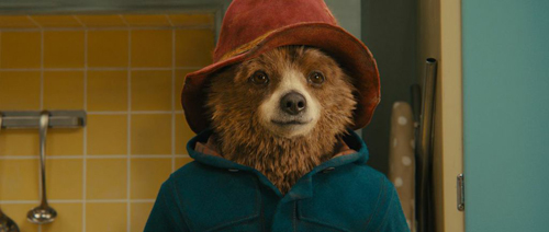 A brown bear wearing a red hat and a teal jacket standing in a room with yellow tiles.