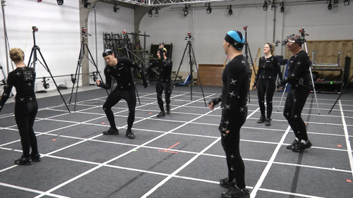 Five people in black motion capture suits stand in a studio with cameras on tripods.