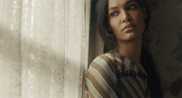 A woman in a striped shirt gazes thoughtfully out of a window through sheer curtains.