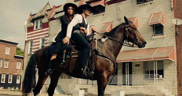 A person in a black hat and dress sits behind another person in a white shirt and black hat on a brown horse.