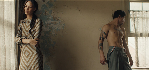 A woman in a striped dress stands by a curtain while a shirtless man stands near a window in a room with peeling paint.