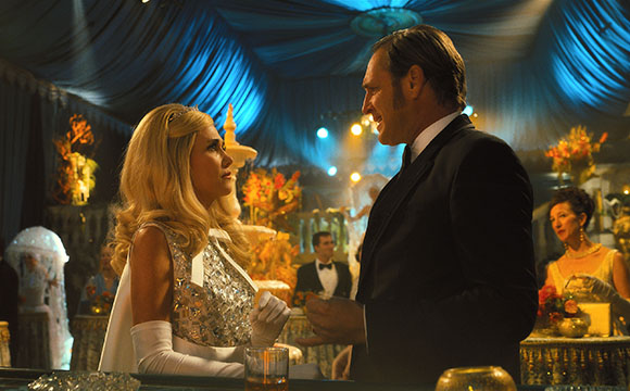 A man in a tuxedo and a blonde woman in a silver gown talk in a blue draped tent.