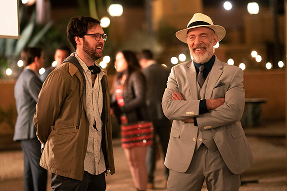 A man in a brown jacket and a man in a grey suit and fedora smile while talking at an outdoor evening event.