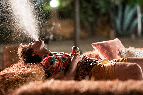 A person in a patterned shirt lies on hay at night, spraying a mist of liquid from their mouth.