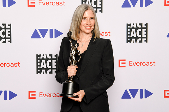 A woman with long blonde hair wearing a black blazer holds a silver trophy against a branded backdrop.
