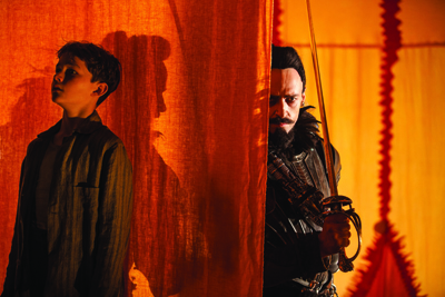 A young boy looks upward while a man with a mustache holds a sword behind an orange fabric curtain.