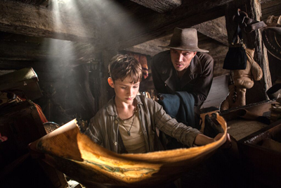 A man in a fedora and a young boy look down at a large, weathered parchment in a dark, dusty attic.
