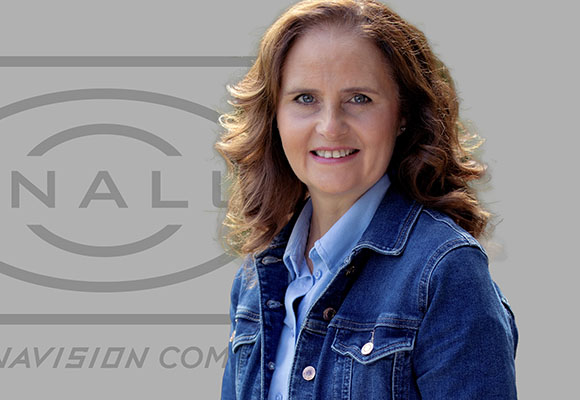 A smiling woman with wavy brown hair wearing a blue denim jacket over a light blue shirt.