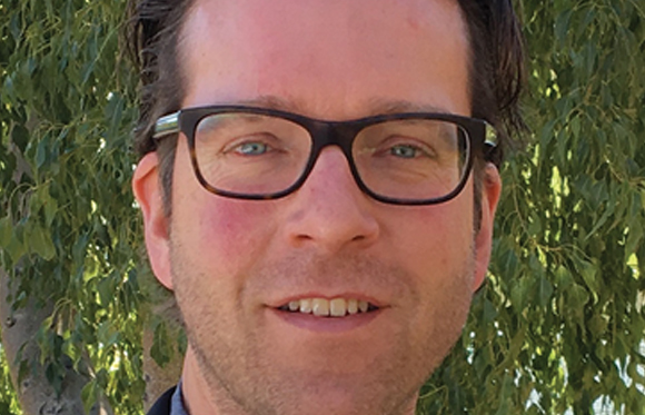 A man with dark hair and black-rimmed glasses smiling in front of green leafy trees.