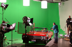 Film crew filming a red convertible car in a studio with a large green screen background.