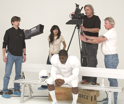 A film crew with a camera and lighting equipment filming a man in white football uniform sitting on a bench.