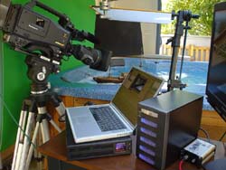 A professional video camera on a tripod, a laptop, and electronic recording equipment on a wooden desk.