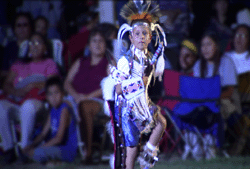 A young person in elaborate feathered regalia performs a traditional dance before an audience.