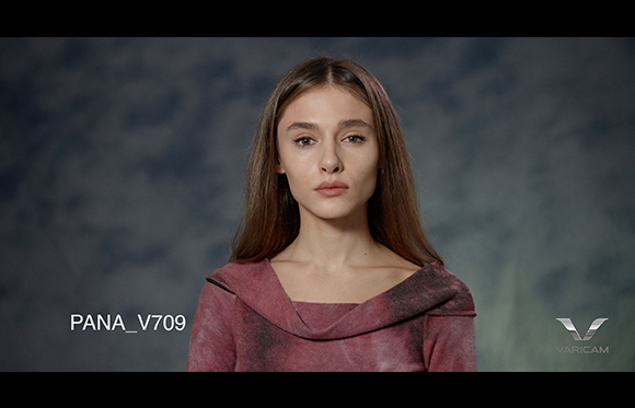 A woman with long brown hair and a pink top looks forward against a dark, textured background.