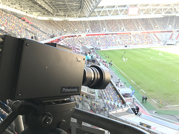 A black Panasonic video camera on a tripod overlooking a green soccer field and stadium seating.
