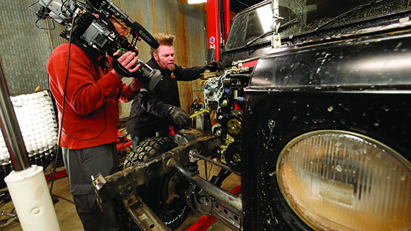 A cameraman films a man working on the engine of a black vehicle in a garage.