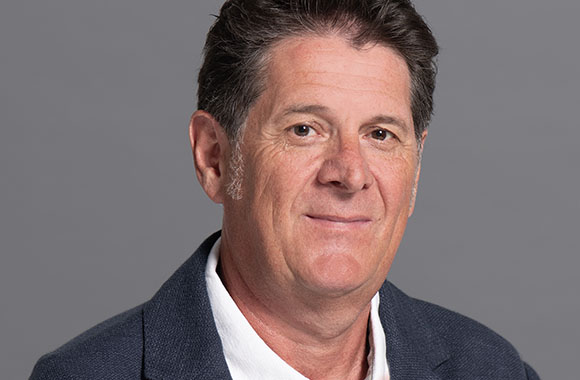A middle-aged man with dark hair wearing a dark blazer and a white collared shirt against a gray background.