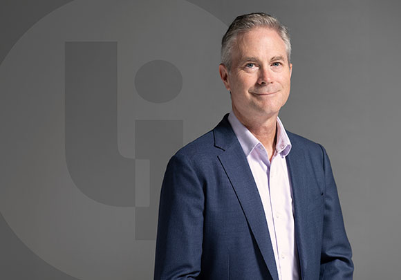 A middle-aged man with graying hair wearing a blue blazer and pink shirt, smiling against a gray backdrop.