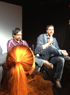 Two men sit on a stage, one speaking into a microphone. A person with red hair is in the foreground.