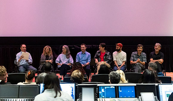 Seven people sit in a row on a stage in front of a large blank screen during a panel discussion.