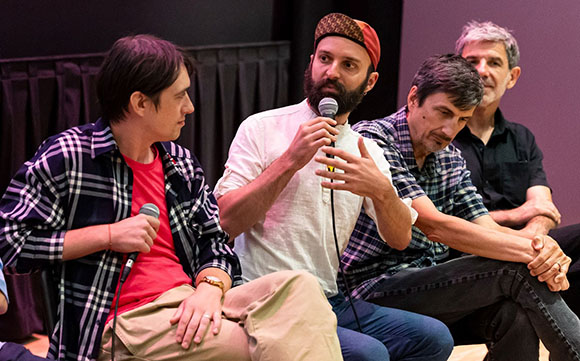 Four men sit in a row on a stage, one man in a white shirt speaks into a microphone.