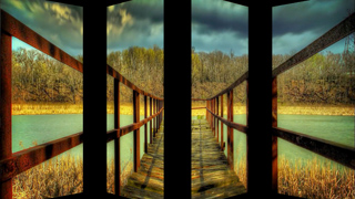 A wooden boardwalk with metal railings leads toward a line of trees across a body of water under dark clouds.
