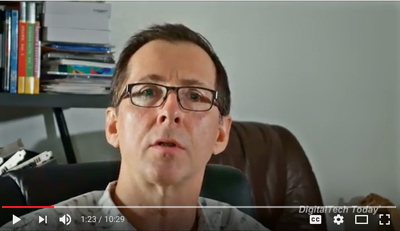 A man with short dark hair and glasses looks forward against a plain wall and bookshelf background.