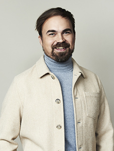 A smiling man with dark hair and a beard wearing a light beige jacket over a blue turtleneck.