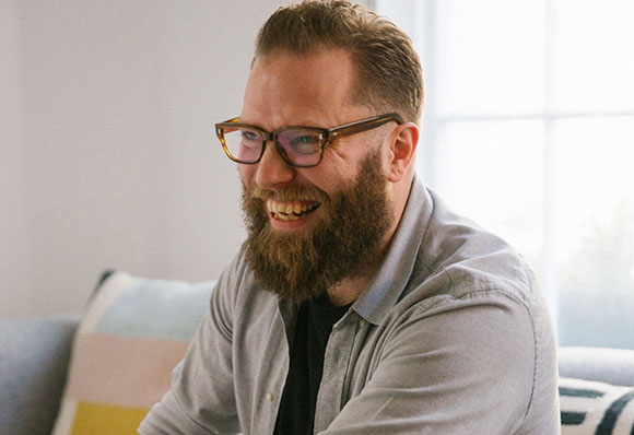A bearded man wearing glasses and a grey button-down shirt smiles while looking to the side.