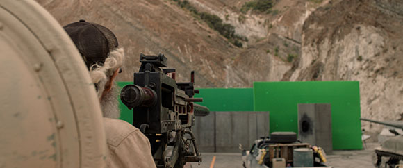 A man in a cap aims a large machine gun at a green screen set within a rocky quarry landscape.