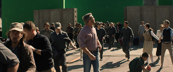 A crowd of people standing in an open area with a large green screen behind them.