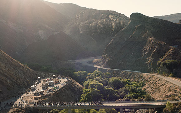 A high-angle view of a winding road through a rugged, sunlit canyon with green trees and steep hills.