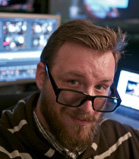 A man with a beard and black-rimmed glasses looks toward the camera in a dimly lit room with computer monitors.