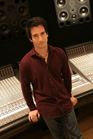 A man in a maroon shirt stands beside a large audio mixing console in a studio.