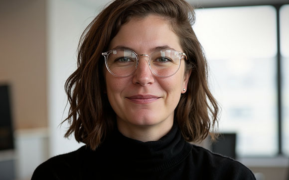 A woman with wavy brown hair and round glasses smiling in an office setting.