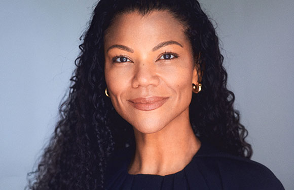 A smiling woman with long, dark curly hair and gold hoop earrings against a gray background.